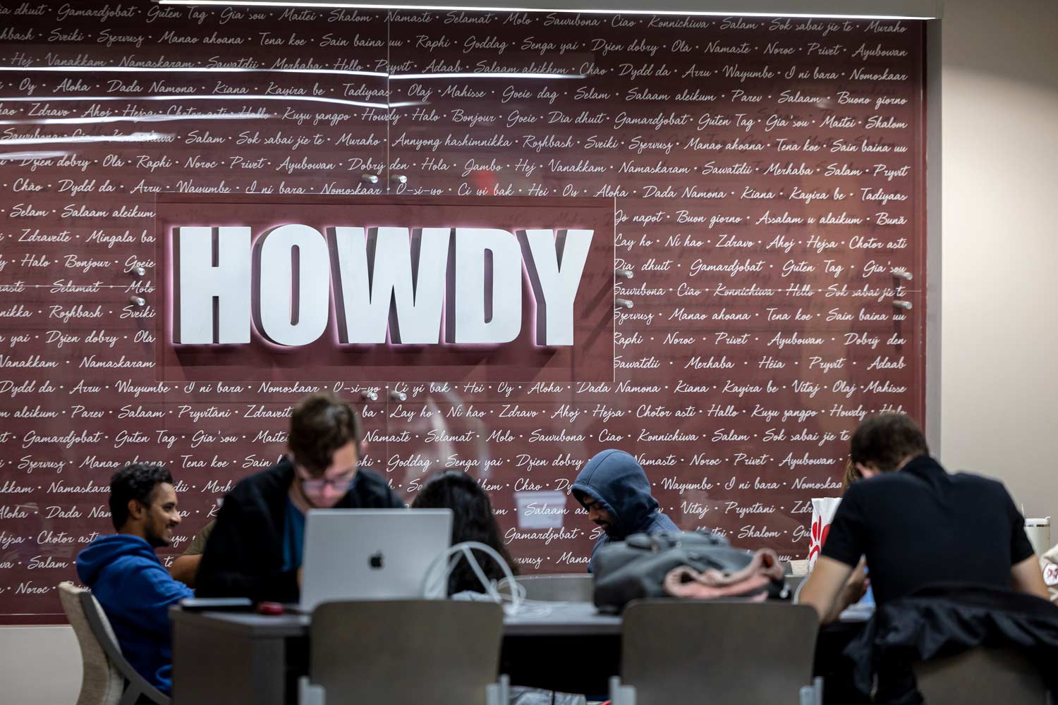 Students work diligently on their laptops in front of a lit up sign that says HOWDY