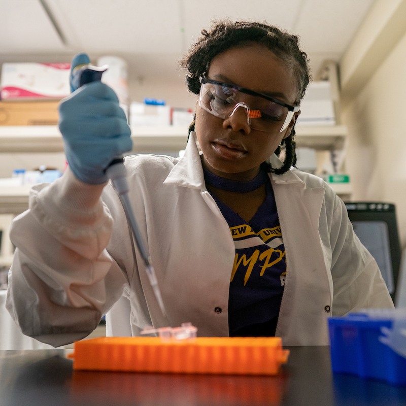 A student wearing a lab coat and protective goggles works with chemicals in a lab